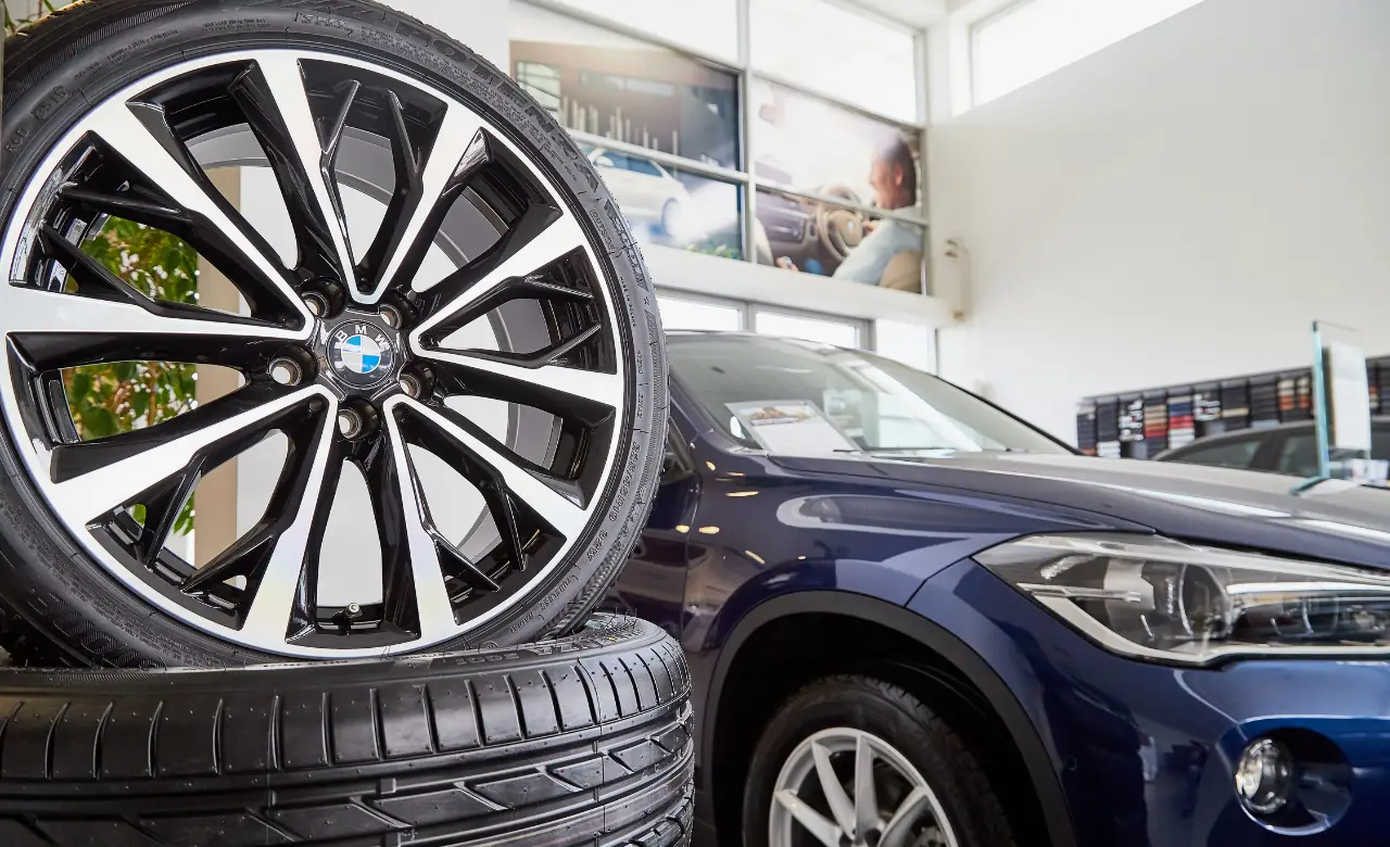 Close-up of a car showroom featuring a black and silver wheel rim with a blue and white logo, stacked on tires. In the background, a blue SUV is parked under bright lighting with large windows overhead. EuroMechanic | McDermott Motors Auto | Top Reviewed Auto Repair Shop in Toronto | Visit mcdermottmotors.com