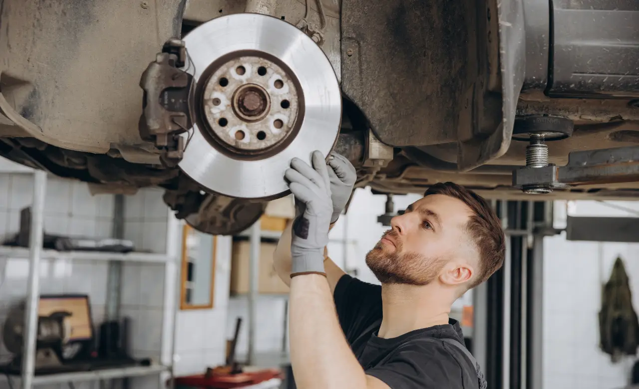 A mechanic wearing gloves inspects a cars brake disc in a garage. The car is lifted on a hydraulic lift, revealing the underside and wheel area. Shelves with tools and equipment are visible in the background. EuroMechanic | McDermott Motors Auto | Top Reviewed Auto Repair Shop in Toronto | Visit mcdermottmotors.com