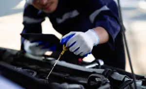 A mechanic in a blue uniform and white gloves checks the oil level of a car engine using a dipstick. The mechanic holds a tablet in one hand and leans over the open car hood, focusing on the task. EuroMechanic | McDermott Motors Auto | Top Reviewed Auto Repair Shop in Toronto | Visit mcdermottmotors.com
