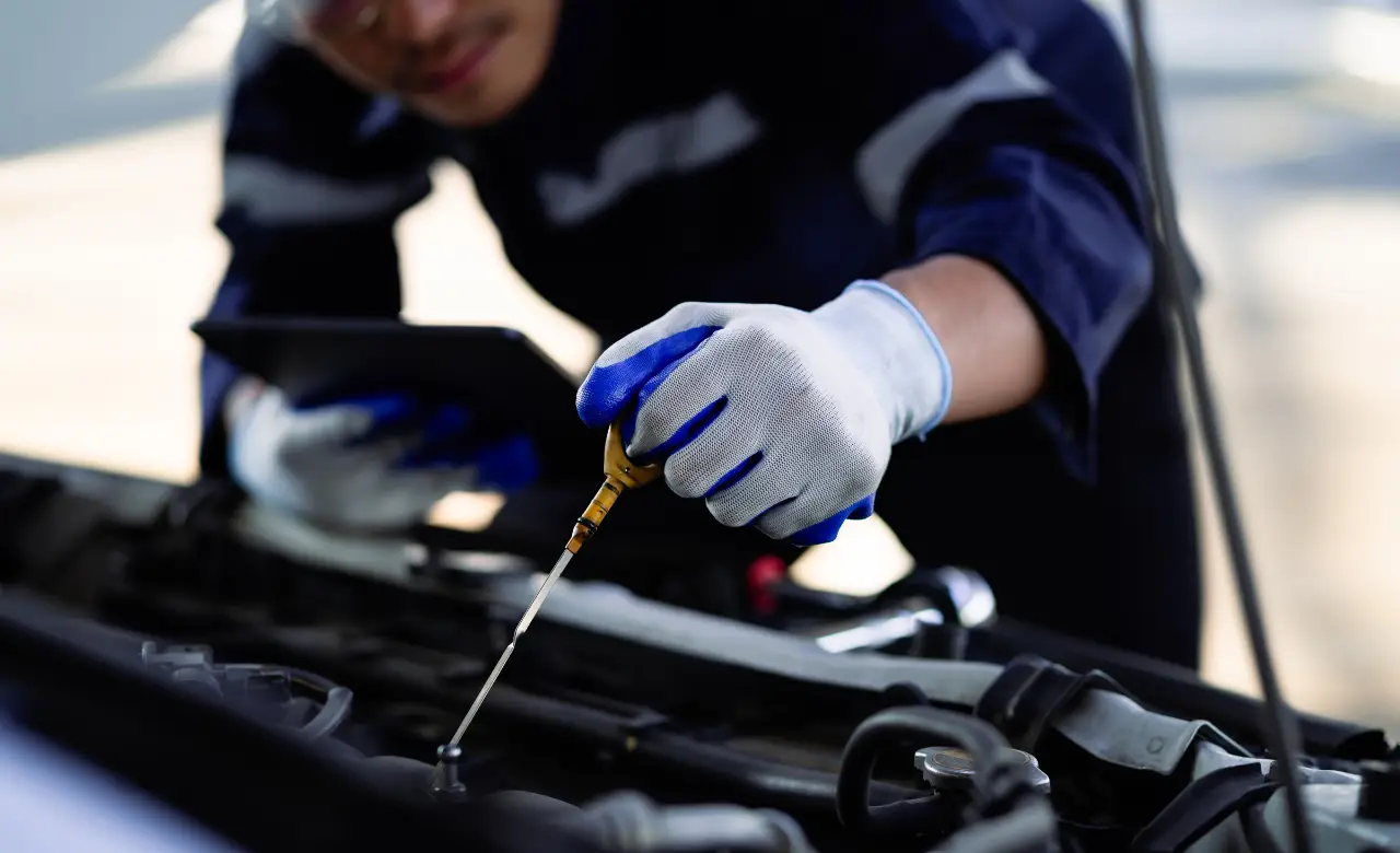 A mechanic in a blue uniform and white gloves checks the oil level of a car engine using a dipstick. The mechanic holds a tablet in one hand and leans over the open car hood, focusing on the task. EuroMechanic | McDermott Motors Auto | Top Reviewed Auto Repair Shop in Toronto | Visit mcdermottmotors.com