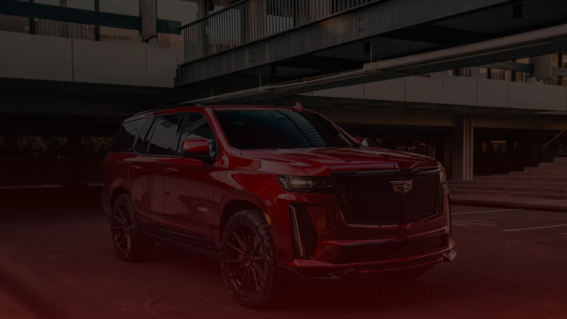 A shiny, red Cadillac SUV is parked in an urban setting, partially illuminated by soft lighting. The vehicle has large black wheels and is positioned at an angle, showcasing its front and side. Dark architectural structures form the backdrop in this Toronto scene. EuroMechanic | McDermott Motors Auto | Top Reviewed Auto Repair Shop in Toronto | Visit mcdermottmotors.com