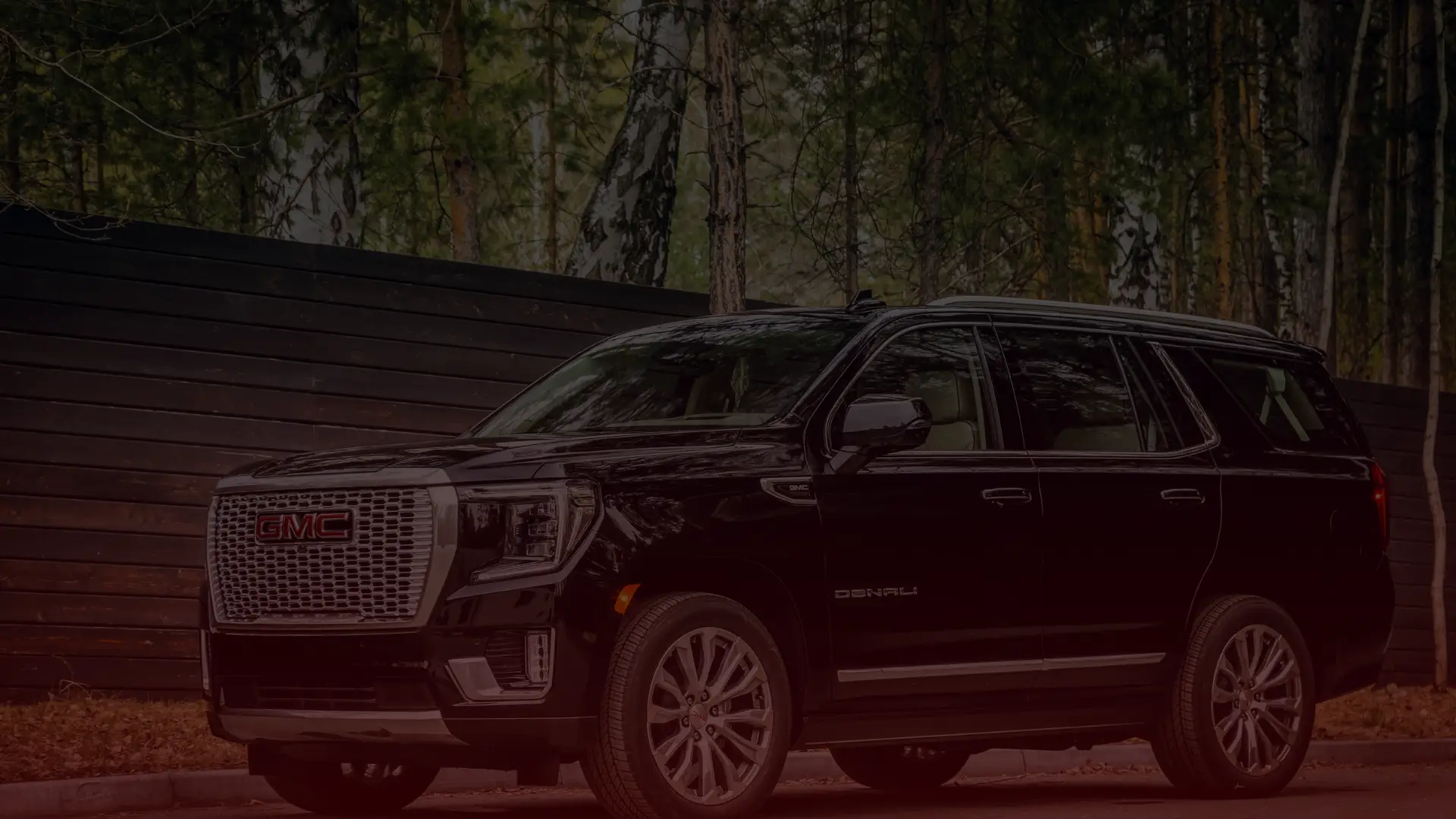 A black GMC Denali SUV is parked on a road with a dark wooden fence and tall trees in the background, as if waiting for its next adventure. The vehicles grille and headlights gleam, a testament to the craftsmanship admired by any GM mechanic in Toronto. EuroMechanic | McDermott Motors Auto | Top Reviewed Auto Repair Shop in Toronto | Visit mcdermottmotors.com