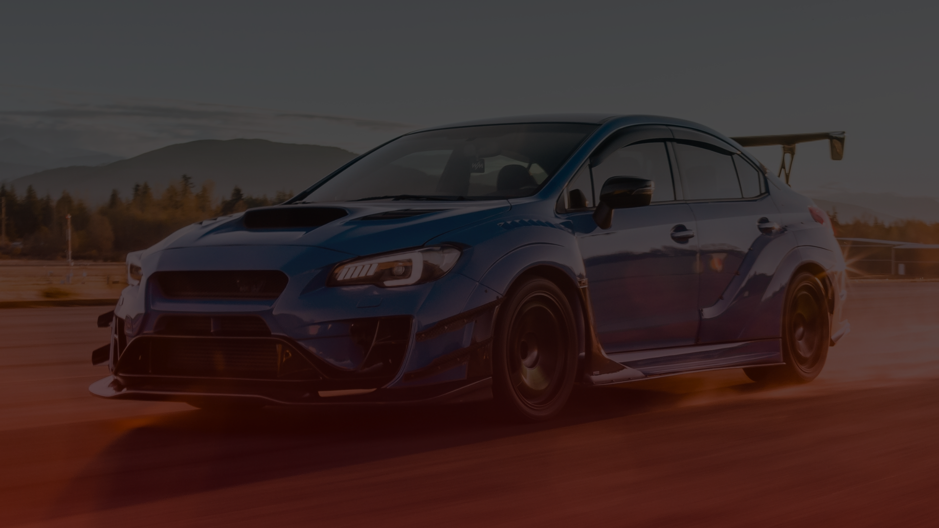 A blue sports car with a large rear spoiler races on an open road at sunset, reminiscent of a Subaru tuned by a top mechanic in Toronto. Its aerodynamic features and sleek design blur into the speed-laden scene, as distant mountains and trees complete the stunning backdrop. EuroMechanic | McDermott Motors Auto | Top Reviewed Auto Repair Shop in Toronto | Visit mcdermottmotors.com