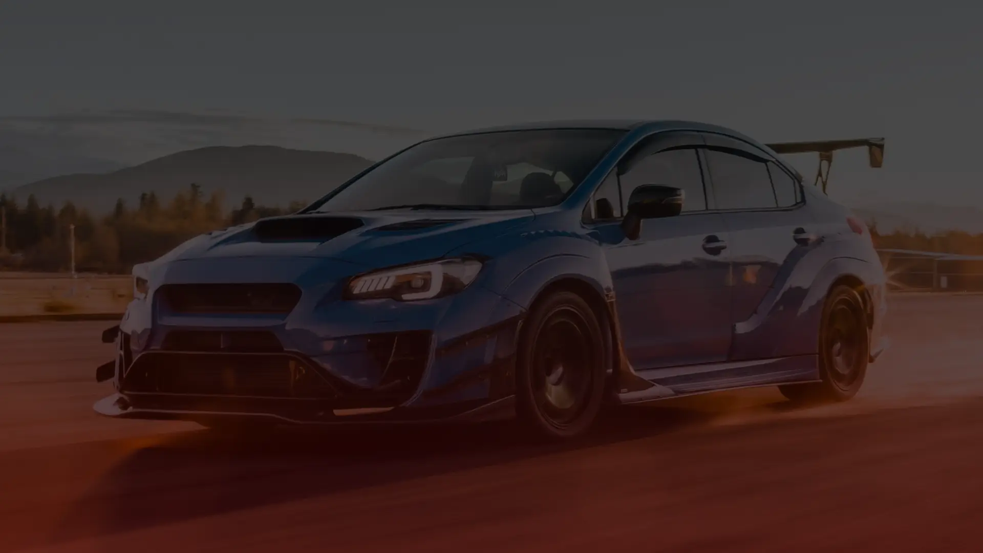 A blue sports car with a large rear spoiler races on an open road at sunset, reminiscent of a Subaru tuned by a top mechanic in Toronto. Its aerodynamic features and sleek design blur into the speed-laden scene, as distant mountains and trees complete the stunning backdrop. EuroMechanic | McDermott Motors Auto | Top Reviewed Auto Repair Shop in Toronto | Visit mcdermottmotors.com