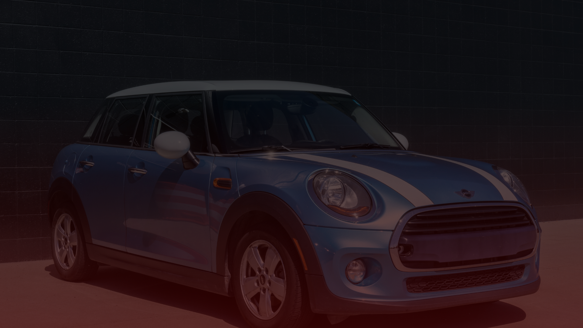 The vibrant Blue Mini Cooper, adorned with white racing stripes, sits elegantly against a dark brick wall in Toronto. Its black trim and alloy wheels complement the white mirrors and roof, all illuminated by the daytime light. EuroMechanic | McDermott Motors Auto | Top Reviewed Auto Repair Shop in Toronto | Visit mcdermottmotors.com