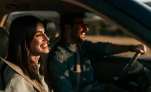 A woman and a man are smiling and sitting in a car, with the man driving. The sunlight creates a warm atmosphere inside the vehicle. Both are wearing casual clothing and seem to be enjoying a pleasant drive. EuroMechanic | McDermott Motors Auto | Top Reviewed Auto Repair Shop in Toronto | Visit mcdermottmotors.com