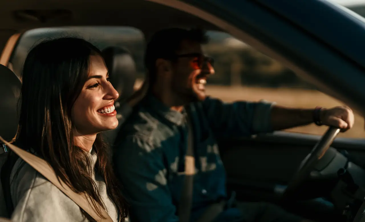 A woman and a man are smiling and sitting in a car, with the man driving. The sunlight creates a warm atmosphere inside the vehicle. Both are wearing casual clothing and seem to be enjoying a pleasant drive. EuroMechanic | McDermott Motors Auto | Top Reviewed Auto Repair Shop in Toronto | Visit mcdermottmotors.com